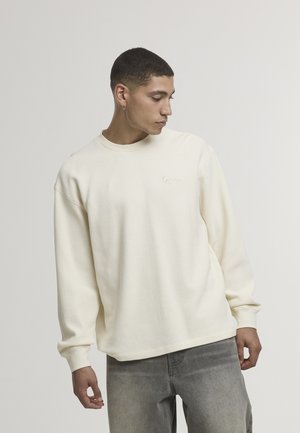 Cremefarbener, strukturierter Sweatshirt mit Rundhalsausschnitt und lockerem Schnitt. Verfügt über gerippte Bündchen und Halsausschnitt, besticktes Logo auf der Brust, getragen mit grauen Jeans.