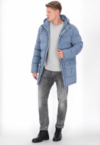 Jasnoniebieska pikowana kurtka z kapturem, zapinana na suwak, z przednią kieszenią. Kurtka ma pikowany wzór i jest zestawiona z szarymi spodniami oraz czarnymi butami.