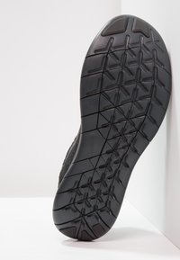 Semelle de chaussure de sport noire avec un motif de bande de roulement texturé. Le matériau semble en caoutchouc, avec un talon incurvé et un design minimaliste.