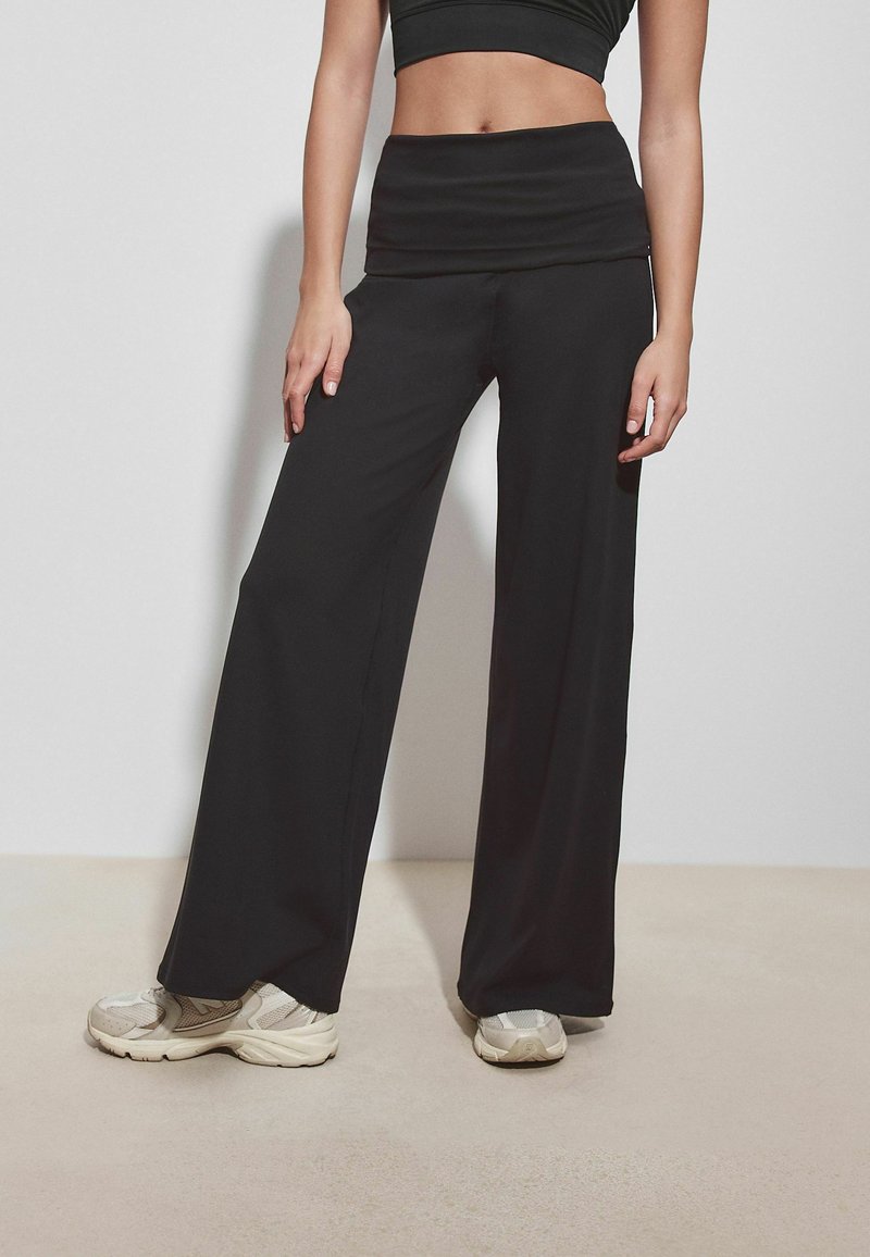 Woman wearing black wide-leg pants and a black crop top, standing on light-colored floor against a plain light wall.