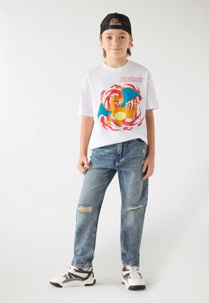 Ragazzo con cappellino nero, maglietta bianca Pokémon con Charizard, jeans blu strappati e sneakers bianche, in piedi su uno sfondo bianco.