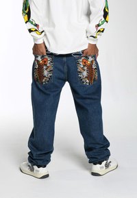 Blue denim pants with large embroidered koi fish on pockets; loose fit, elastic waistband, paired with white sneakers featuring black design accents.