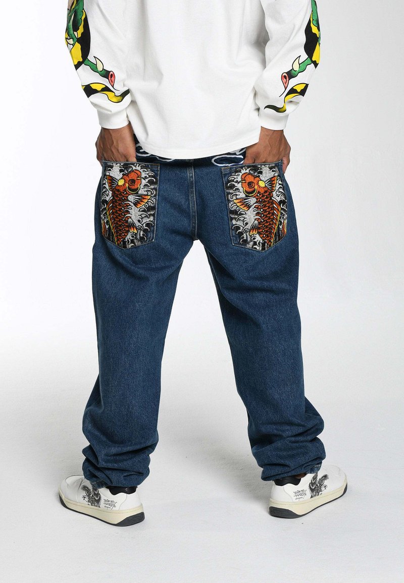 Blue denim pants with large embroidered koi fish on pockets; loose fit, elastic waistband, paired with white sneakers featuring black design accents.