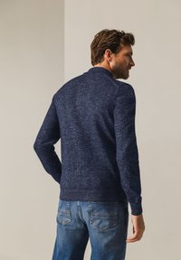 Marineblauwe gebreide trui met een textuurafwerking en ronde halslijn, gecombineerd met lichtblauwe jeans met stikseldetails op de achterzak.