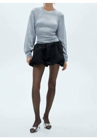 Eine Frau trägt einen hellblauen langärmeligen Pullover, schwarze gepuffte Shorts, transparente schwarze Strumpfhosen und silberne High-Heels mit offenem Zeh.