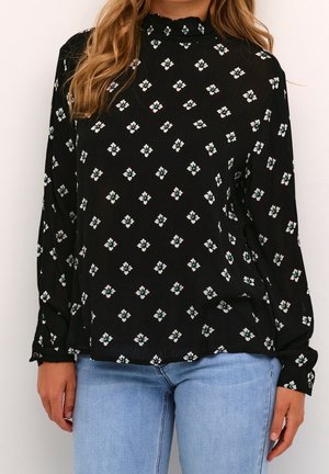 Kvinde iført sort langærmet bluse med lille hvidt blomsterprint og høj krave, kombineret med lyseblå jeans.