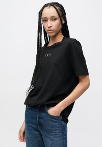 Μαύρο βαμβακερό T-shirt με κοντά μανίκια, με στρογγυλή λαιμόκοψη και μικρή λεπτομέρεια λογότυπου στο στήθος· φορεμένο με μπλε τζιν παντελόνι.