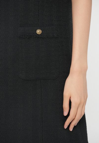 Fekete textúrájú szövetruha, elülső zsebbel, amelynek arany gombja van. Zsúfolt közeli kép a zsebről és a sima felületre tett kézről.