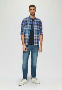 Gepofte blauw en groene shirt over een zwart graphic t-shirt, gecombineerd met lichtblauwe jeans en witte sneakers met beige accenten.