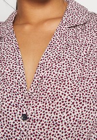 Ljusrosa skjorta med svart och djupröd prickmönster, kragsdesign och en enda svart knapp vid halsen. Slät tygtextur.