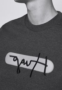 Personne portant un t-shirt à col rond foncé avec une forme ovale blanche et une broderie cursive abstraite noire sur la poitrine.