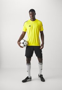 T-shirt de sport jaune avec des accents noirs, shorts noirs, chaussettes blanches avec des rayures noires et chaussures de foot noires, tenant un ballon de football.