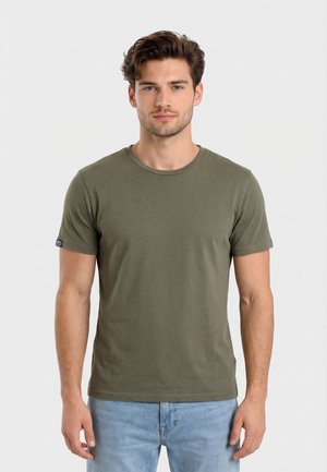 Mand, der står med front mod kameraet iført en olivengrøn kortærmet t-shirt og lyseblå jeans mod en ensfarvet baggrund.