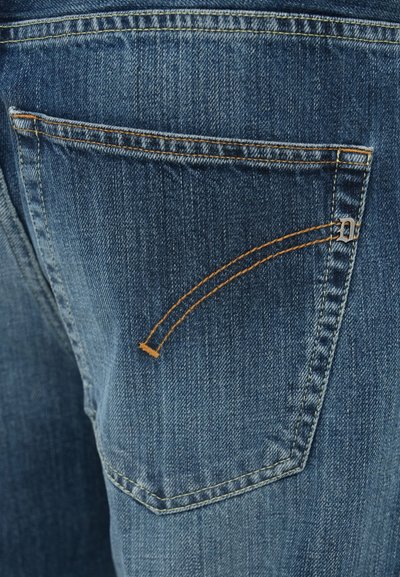 Κοντινή λήψη ύφασματος ντένιμ που δείχνει μια πίσω τσέπη με πορτοκαλί ραφές και μια μικρή ασημένια διακόσμηση με λογότυπο. Η υφή είναι λεία με μια μπλε χρωματική βαθμίδωση.