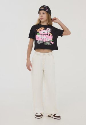 T-shirt nera cropped con grafica testuale colorata, abbinata a pantaloni bianchi a gamba larga e sneaker in bianco e nero. Sfondo semplice.