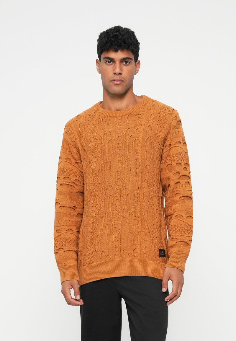 Πορτοκαλί πλεκτός πουλόβερ με υφές από καλώδια, ριγέ μανσέτες και στρίφωμα. Χαλαρή εφαρμογή, συνδυασμένο με σκούρους παντελόνες.