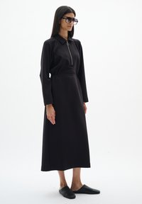 Femme portant une longue robe noire avec une fermeture éclair à l'avant, des chaussures noires sans lacets et de grandes lunettes de soleil foncées, se tenant devant un fond blanc.