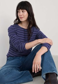 Violetais dryžuotas ilgomis rankovėmis viršus su apvaliu iškirpimu, derinamas su aukštu liemeniu mėlynomis džinsomis. Kasdienis derinys su minkšta tekstūra, dėvimas sėdint modelyje.