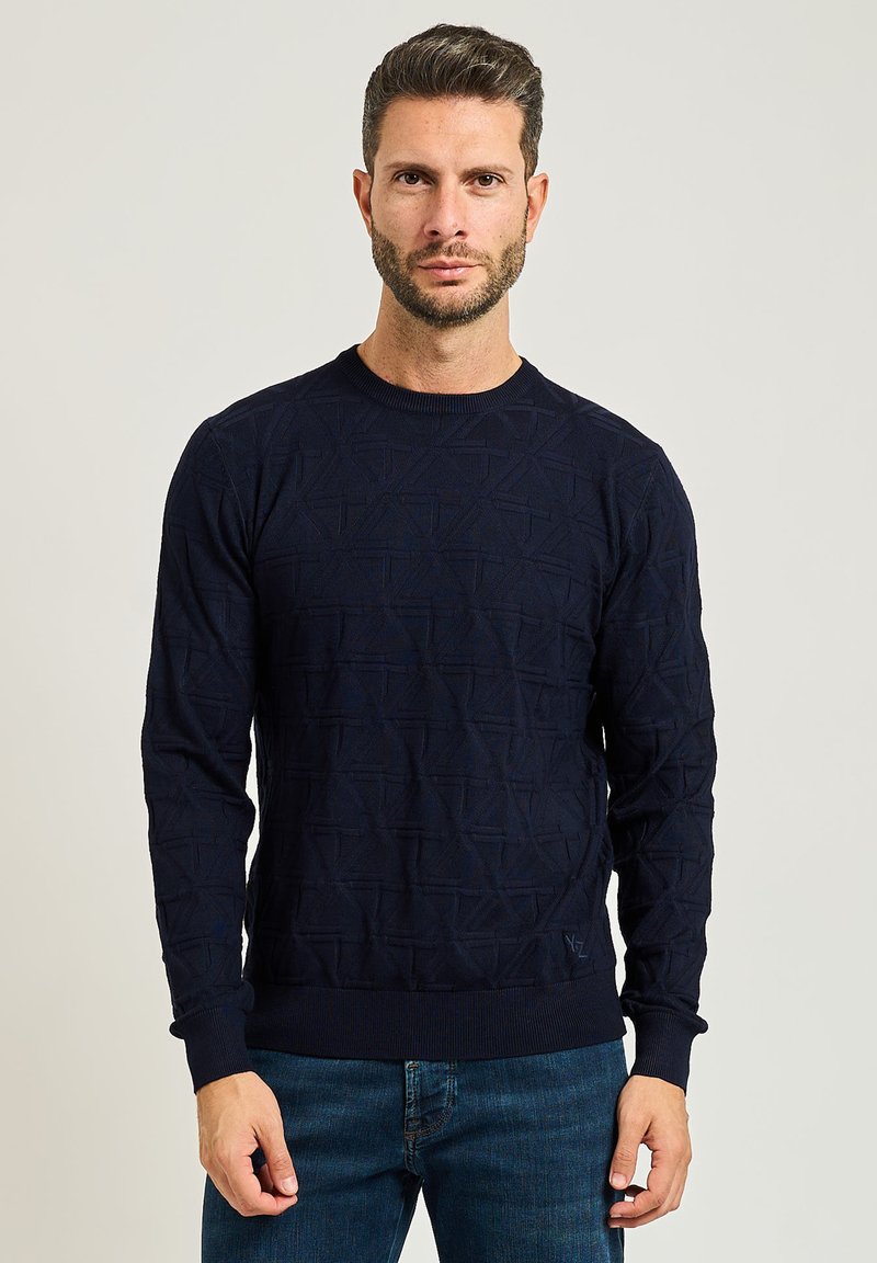 Maglione lavorato a maglia navy con texture geometrica, scollatura a girocollo e polsini a coste. Abbinato a jeans in denim blu, il modello si trova davanti a uno sfondo semplice.