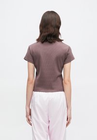 Άτομο με καστανά μακριά μαλλιά που φοράει ένα κοντομάνικο μωβ μπλουζάκι και άνετα ανοιχτό ροζ παντελόνια, στέκοντας μπροστά σε μια απλή λευκή ταπετσαρία.