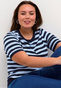 Blå og marine blå stribede kortærmet sweater med rund halsudskæring, der har en tætsiddende pasform. Modellen har denim jeans på og sidder foran en hvid baggrund.