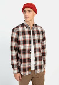 Button-up plaid shirt in gray, red, and black with a collar, long sleeves, and a chest pocket. Worn over a white T-shirt, paired with tan pants.