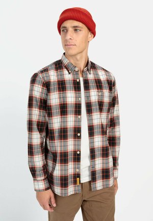 Button-up plaid shirt in gray, red, and black with a collar, long sleeves, and a chest pocket. Worn over a white T-shirt, paired with tan pants.