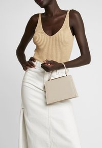 Beżowy dzianinowy top bez rękawów, biały dżinsowy spódniczka z bocznym rozcięciem, trzymająca małą beżową prostokątną torebkę podręczną z górnym uchwytem.