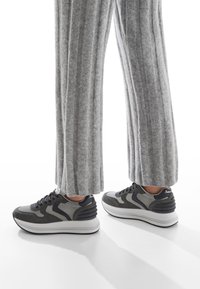 Sneakers grigi con una toma alta testurizzata, accenti neri e grigi e una suola spessa bianca. Indossati con pantaloni lunghi grigi a coste.