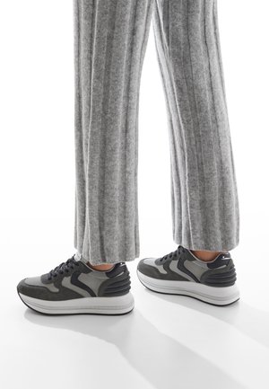 Sneakers grigi con una toma alta testurizzata, accenti neri e grigi e una suola spessa bianca. Indossati con pantaloni lunghi grigi a coste.