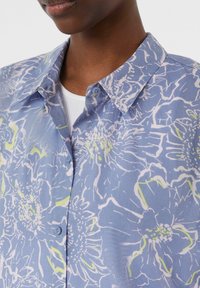 Floral-patterned shirt in light blue, featuring white and green accents. Button-down collar and smooth fabric texture. Close-up view.