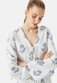 Weißes, leichtes Blusenoberteil aus Stoff mit blauen und braunen floralen Stickereien, V-Ausschnitt und langen, glockenförmigen Ärmeln mit einer Bindegarnitur vorne.