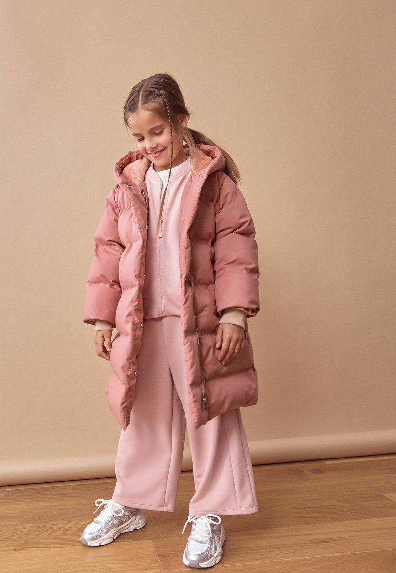 Rosa Puffermantel mit Kapuze, ausgestattet mit einem weichen Innenfutter, getragen über einem hellrosa Sweatshirt und weit geschnittenen Hosen, kombiniert mit silbernen Sneakers.