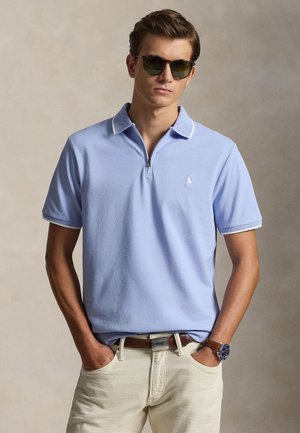 Νέος άνδρας που φοράει γυαλιά ηλίου, ανοιχτό μπλε πόλο με φερμουάρ κοντό μανίκι, μπεζ παντελόνι, καφέ ζώνη και ρολόι χειρός, ποζάροντας με τα χέρια στις τσέπες.