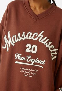 Sweat-shirt marron oversize avec col en V, arborant un texte blanc : "Massachusetts 20 New England" et des détails graphiques supplémentaires. Texture de tissu douce.
