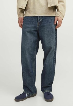 Personne portant un jean bleu foncé ample, une chemise beige à manches longues et des baskets bleu marine, debout sur un fond clair.