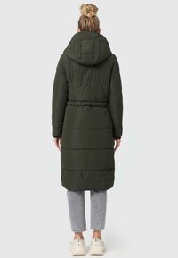 Lange olijfgroene pufferjas met een capuchon, geborduurd ontwerp, taillerende taille en elastische manchetten, gecombineerd met lichtgrijze jeans en witte sneakers.
