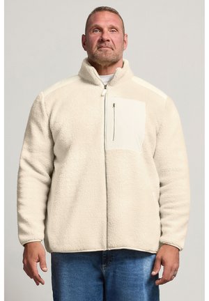 Homme portant une veste polaire crème à fermeture éclair intégrale avec une poche poitrine zippée et un jean bleu, debout devant un fond uni.