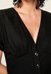 Czarna lniana sukienka z dekoltem w kształcie litery V, krótkimi rękawami, marszczonym detalem przy biuście oraz czterema okrągłymi czarnymi guzikami z przodu.