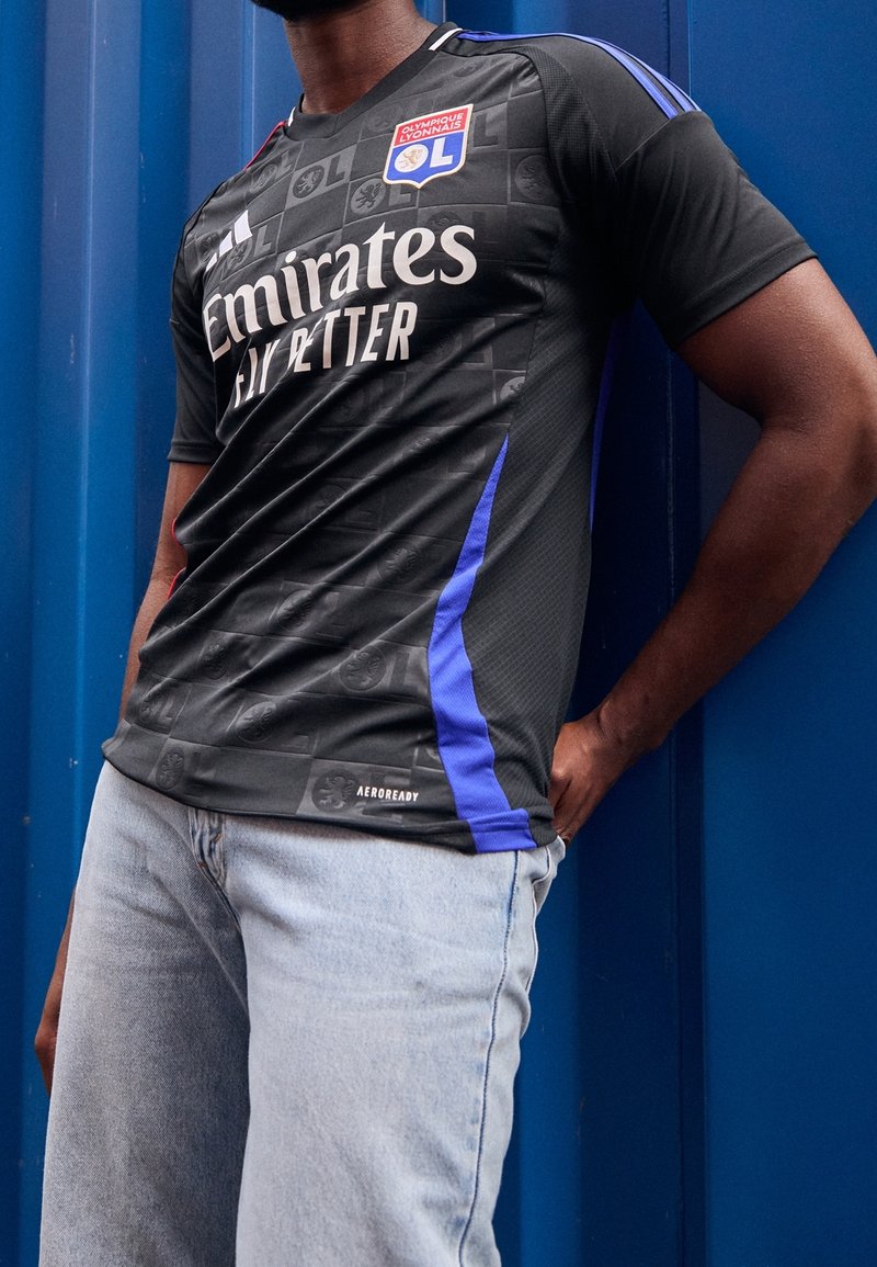 Homme portant un maillot de football noir de l'Olympique Lyonnais avec des accents bleus et un jean bleu clair, debout contre un mur bleu.