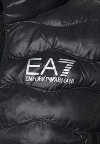 Svart quiltat tyg med vit EA7 Emporio Armani-logotyp och dragkedja på vänster sida.