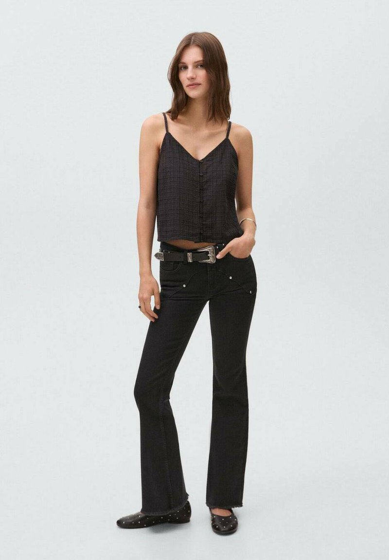 Camiseta negra corta con botones, combinada con jeans negros acampanados que tienen bolsillos delanteros y remaches decorativos. Se lleva con zapatos planos negros.