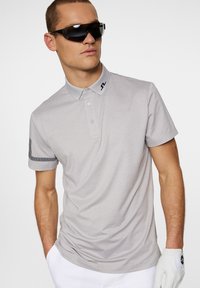 Light gray polo shirt with a collar, three-button placket, and black accent on the sleeve. Worn with black sunglasses and a white glove.