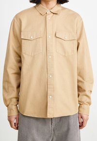 Vaaleanruskea napitettava paita, jossa on klassinen kaulus, kaksi rintataskua ja nepparit. Valmistettu sileästä kankaasta, rento istuvuus.