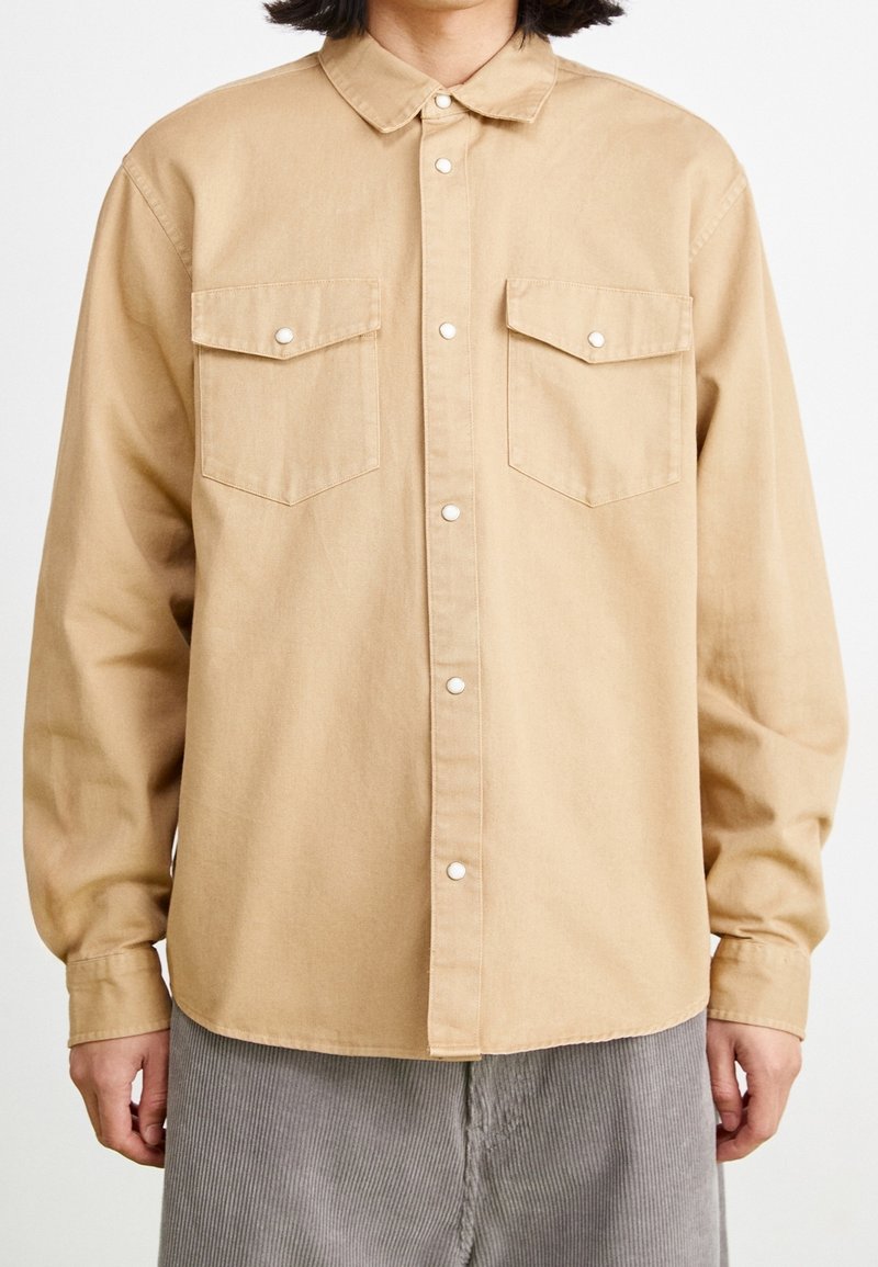 Vaaleanruskea napitettava paita, jossa on klassinen kaulus, kaksi rintataskua ja nepparit. Valmistettu sileästä kankaasta, rento istuvuus.