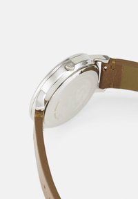 Reloj de acero inoxidable plateado con correa de cuero marrón. Presenta una caja redonda y una corona en el lateral, mostrando un diseño minimalista.