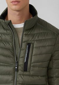 Olivgrüne, gesteppte Jacke mit hohem Kragen, Reißverschluss und einer Brusttasche mit schwarzem Akzent. Glatte Textur mit gestepptem Design.