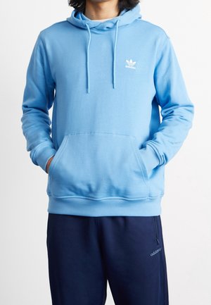 Osoba ubrana w jasnoniebieską bluzę Adidas z kapturem i kieszenią z przodu oraz granatowe spodnie dresowe Adidas, stojąca z rękami w kieszeniach.