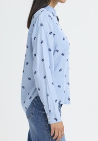 Kvinne i lys blå knappet skjorte med mørkeblå bladmønster og blå jeans, vist fra siden mot en hvit bakgrunn.