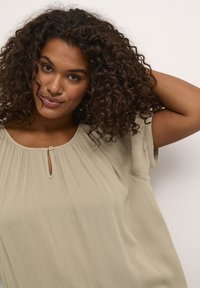Top beige chiaro con un tessuto texturizzato, maniche corte e un'apertura a forma di chiave sul collo. Design fluido e drappeggiato con pieghe.